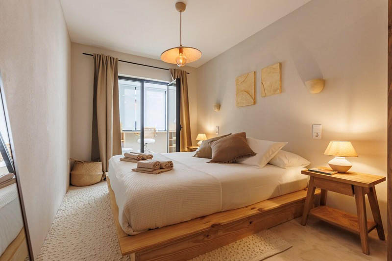 Phòng ngủ homestay tối giản với giường gỗ, ga trắng, tab đầu giường và đèn ngủ ánh sáng vàng.
